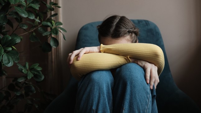 Upset depressed teenager crying at home, sad frustrated teen girl sitting by window with head on knees. Teenagers and mental health concept