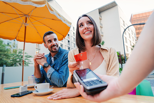 Happy couple paying the bill of the coffee shop using a contactless creditcard payment. Young adult customer woman doing a purchase on a restaurant to the cashier with a debit card. People spending
