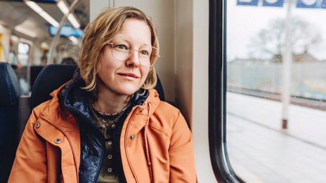A blond woman in an orange jacket sits in a train. She is looking out the window. Outside is a cold rainy autumn day.