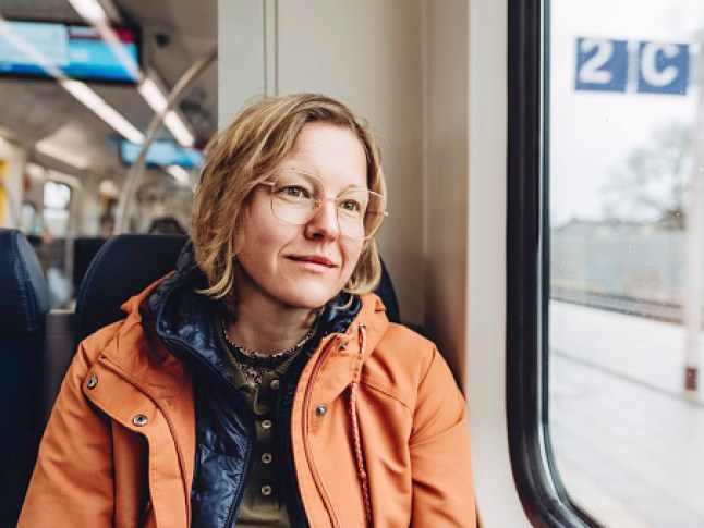 A blond woman in an orange jacket sits in a train. She is looking out the window. Outside is a cold rainy autumn day.