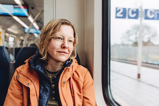 A blond woman in an orange jacket sits in a train. She is looking out the window. Outside is a cold rainy autumn day.