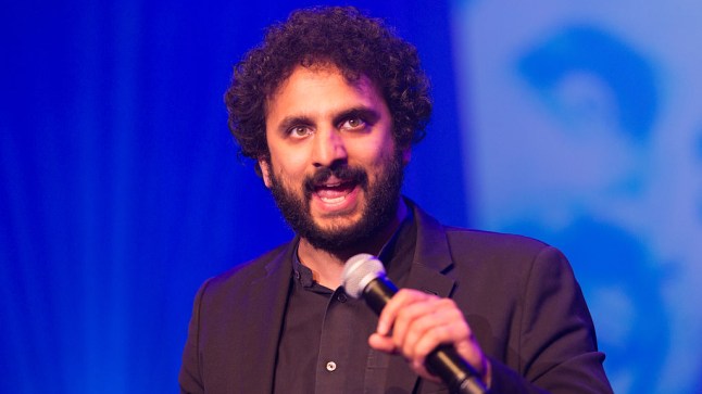 Nish Kumar speaking into microphone on stage while wearing a black suit.