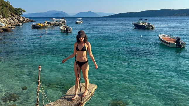 A woman stands on a ledge over blue ocean with boats in the background