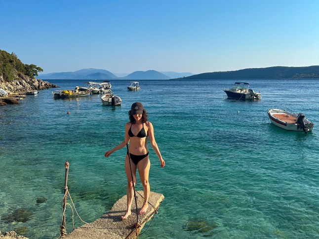 A woman stands on a ledge over blue ocean with boats in the background