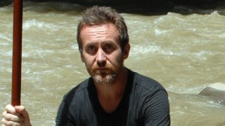 Bruce Parry in Borneo with the Penan tribe holding a stick and sat near a river in BBC's Tribe