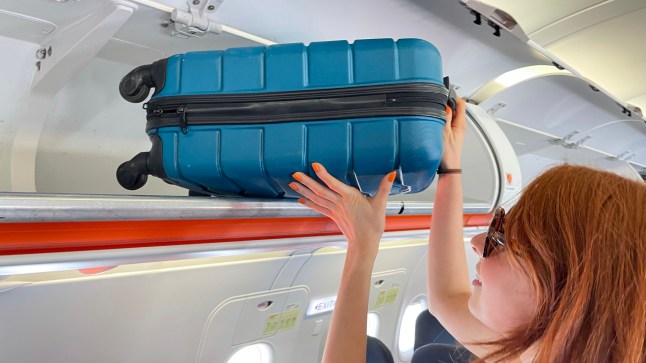 A woman putting her carry on suitcase into an overhead locker on a plane.