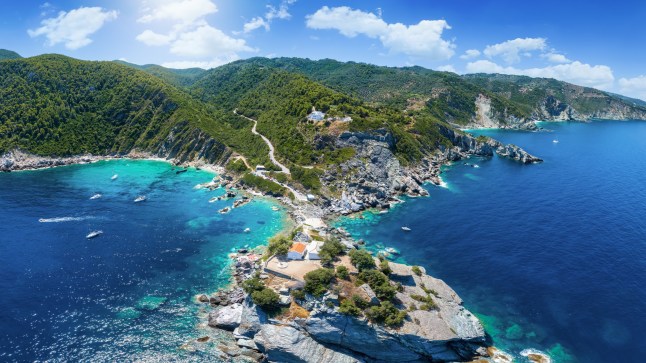 Panoramic aerial view of the coast of Skopelos, and island in Greece, with boats on the water and a winding road into the mountains.