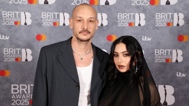 George Daniel in a pinstripe grey jacket and Charli XCX in a black sheer dress with headcover accessory attend The BRIT Awards 2025