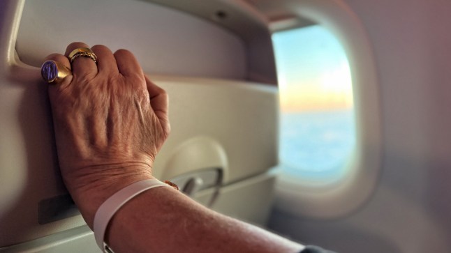Close up of a woman's hand gripping onto a plane seat with a view of the sky through the window in the background.