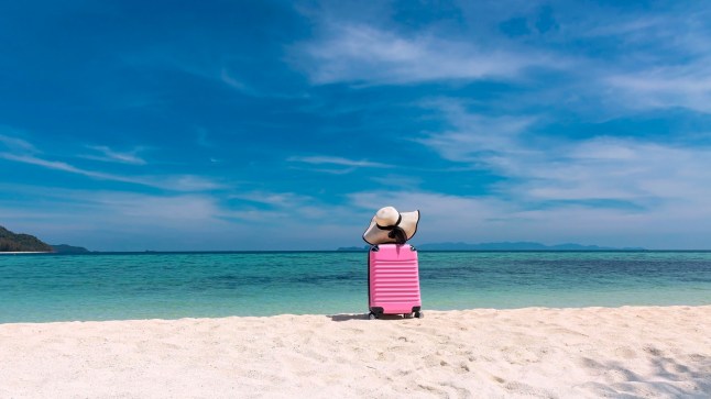 A pink suitcase with a white sunhat on top on a white sandy beach under a blue sky.