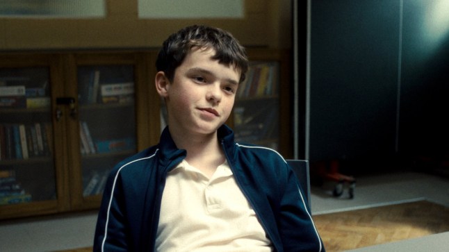 Owen Cooper as Jamie Miller in Adolescence. He is sat behind a table at a detention facility, looking serious.