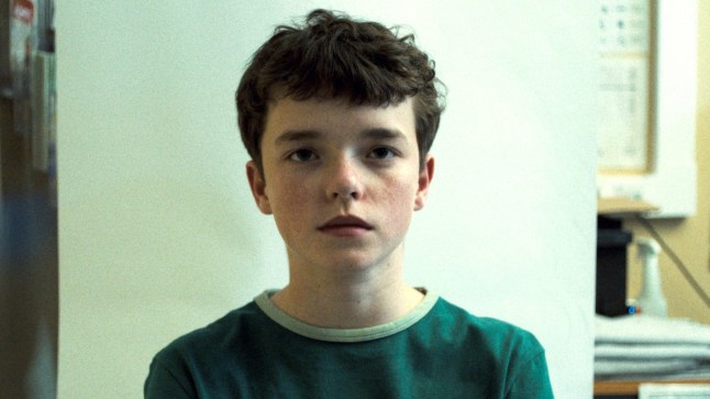 Owen Cooper as Jamie Miller in Adolescence having a photo taken at a police station. He's wearing a green T-shirt