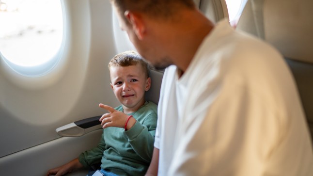 A young boy crying while sat next to a plane window, with his father leaning over him.