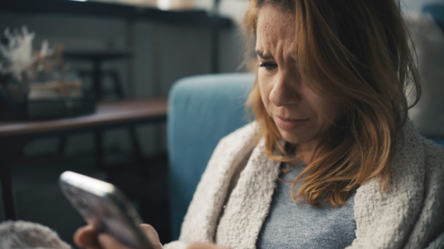 A melancholic mid-adult Caucasian woman sits on the floor, attire casual, absorbed in her phone within the confines of a cozy living room setting.