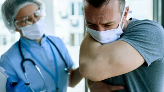 Young man having COVID-19 symptoms and coughing into elbow while talking to doctor at medical clinic.