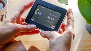 An overhead view of hands holding a home smart meter against a sunlit desk background and checking energy consumption